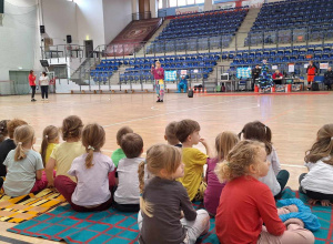 Dzieci siedzą na hali sportowej tyłem do zdjęcia